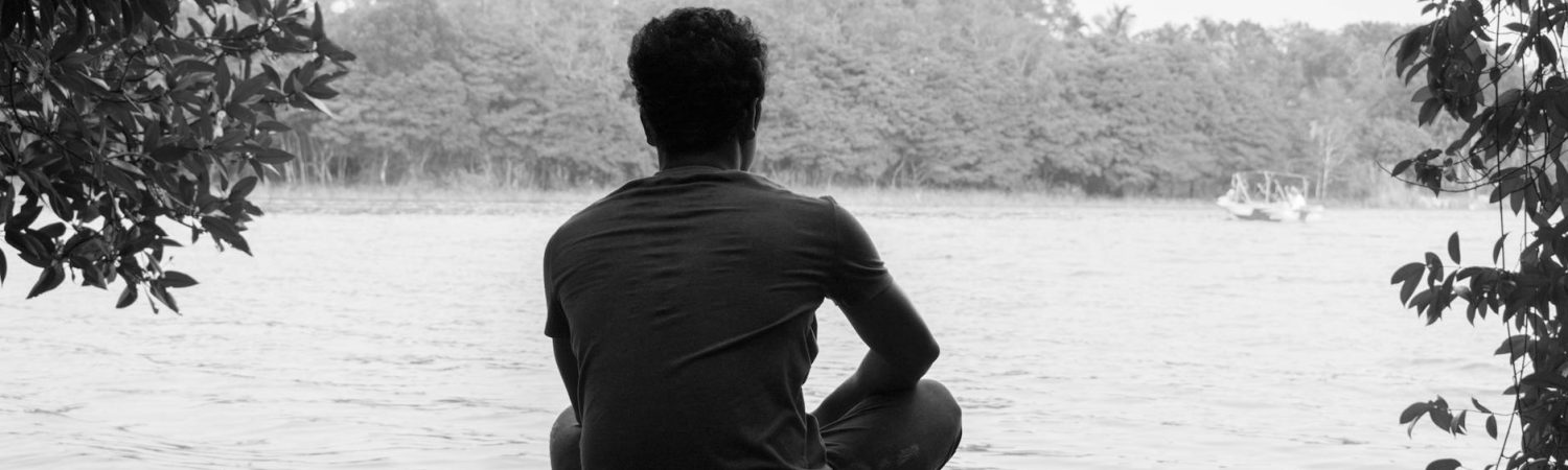 gray scale photography of man sitting on brown wooden floor beside body of water